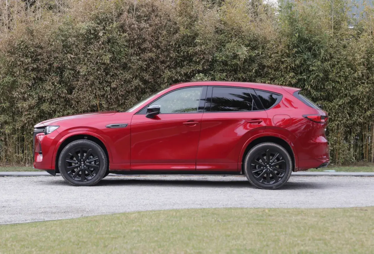 Czerwona Mazda CX-60 3.3 diesel z czarnymi felgami stoi na żwirowej drodze, za nią gęsty żywopłot.