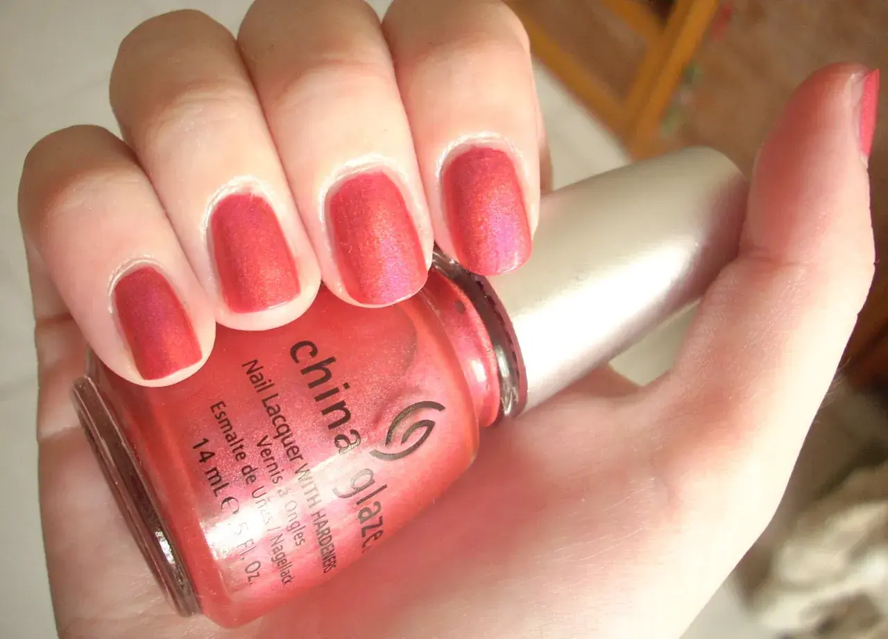 En hand med naglar målade i en glittrig, korallröd nyans. En flaska China Glaze nagellack syns i förgrunden.