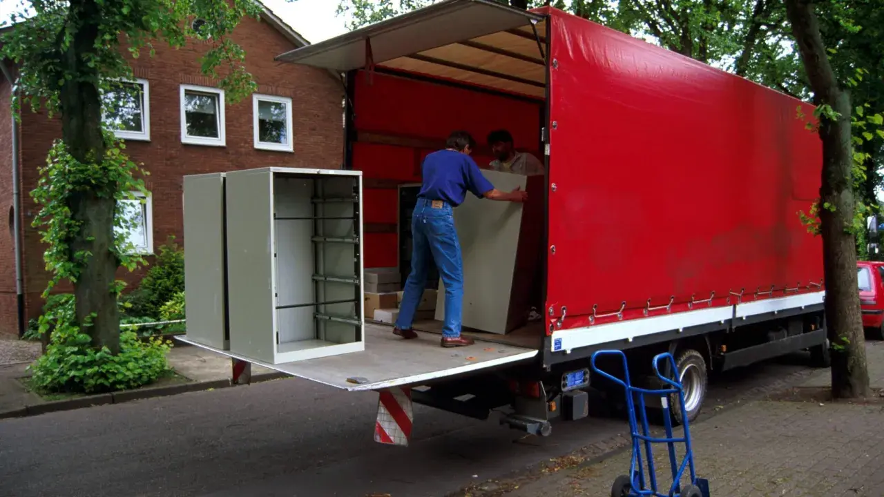 Umzugshelfer laden einen Schrank auf einen roten LKW. Der Schrank steht am Straßenrand, was die Bedeutung von "kerbside" verdeutlicht.