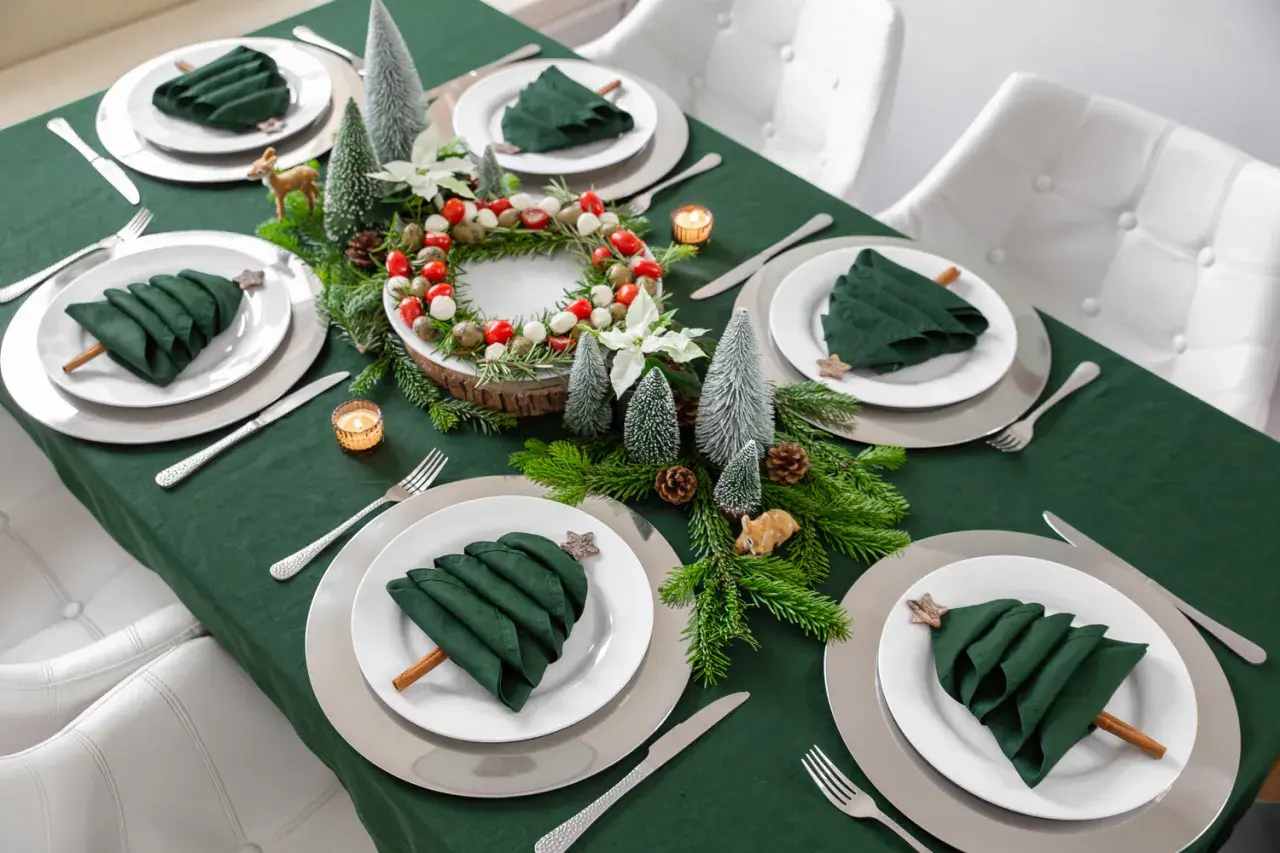 Moderne tischdeko weihnachten: grüne Servietten als Bäume, Tannenzweige, Kerzen und ein Kranz mit Beeren auf einem grünen Tischtuch.