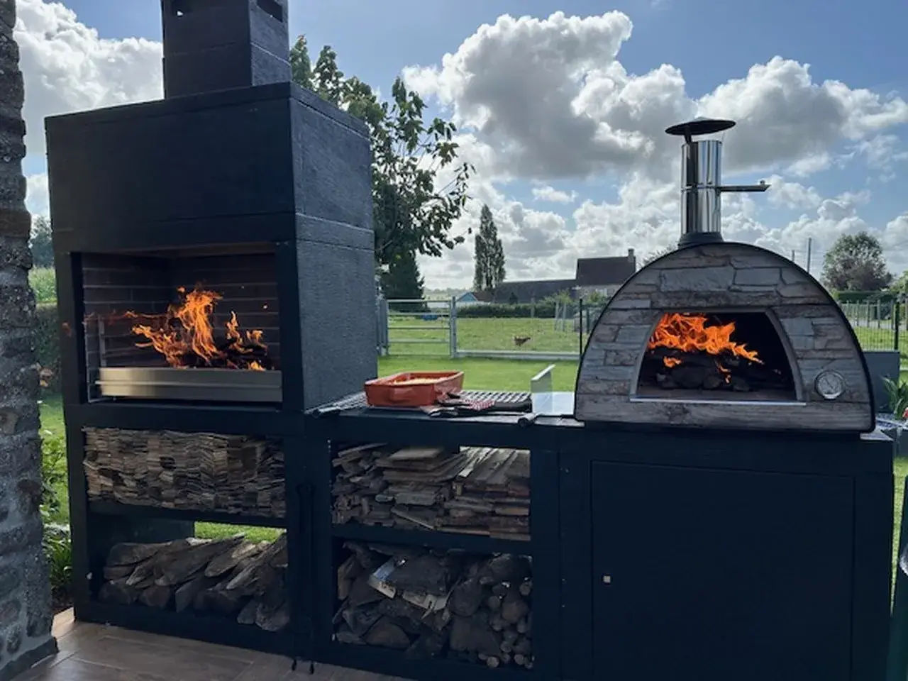 Un grand barbecue et un four à pizza en pierre s'apprêtent à faire un barbecue sous un ciel bleu et nuageux.