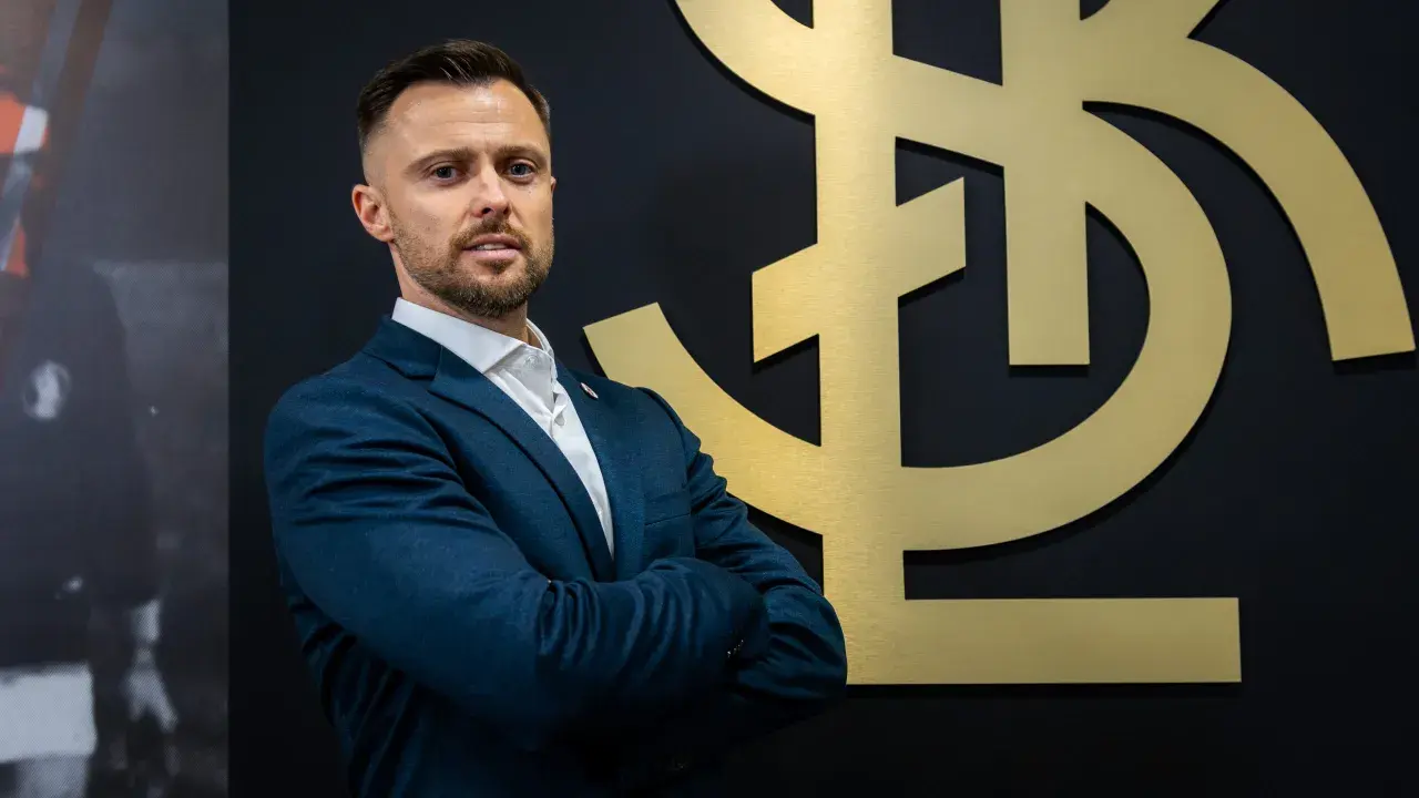 Mężczyzna w garniturze, trener ŁKS Łódź, stoi przed złotym logo klubu.