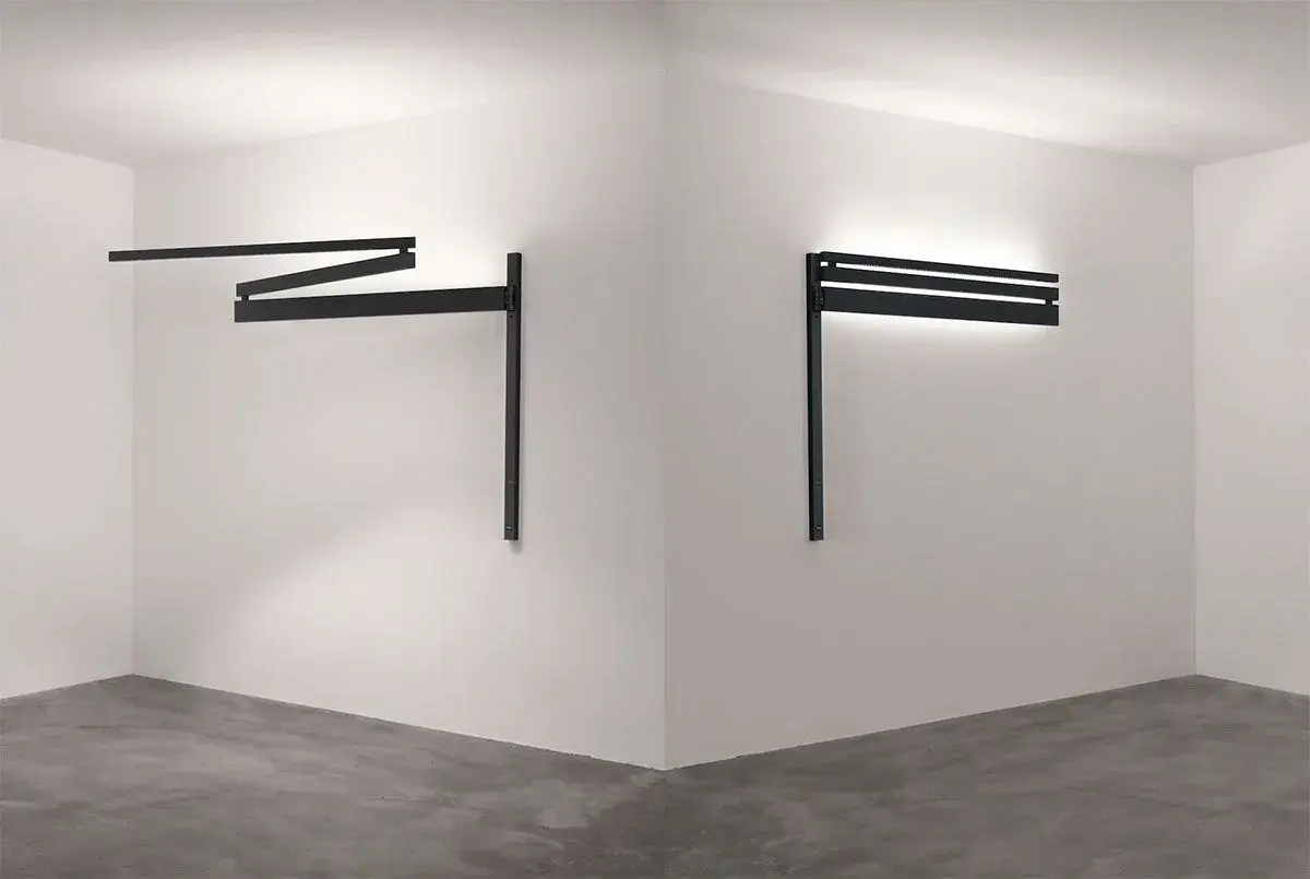 Dos apliques de pared regulables negros, uno extendido en zigzag y otro horizontal con luz indirecta, iluminan un rincón minimalista.