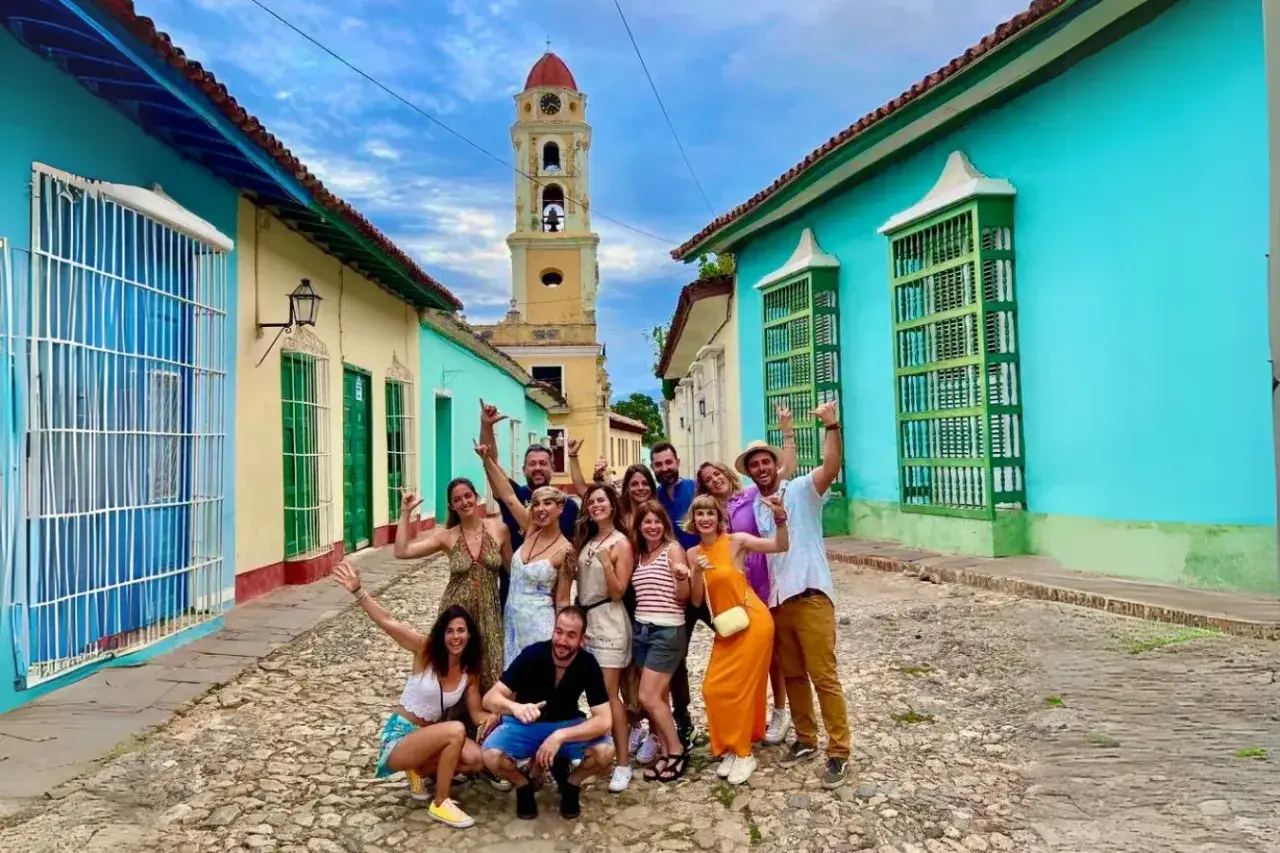 Grupo de amigos sonríe y saluda en una calle empedrada de Trinidad, Cuba, con casas coloridas y una torre de iglesia al fondo. ¡Es seguro viajar a Cuba y disfrutar de su encanto!