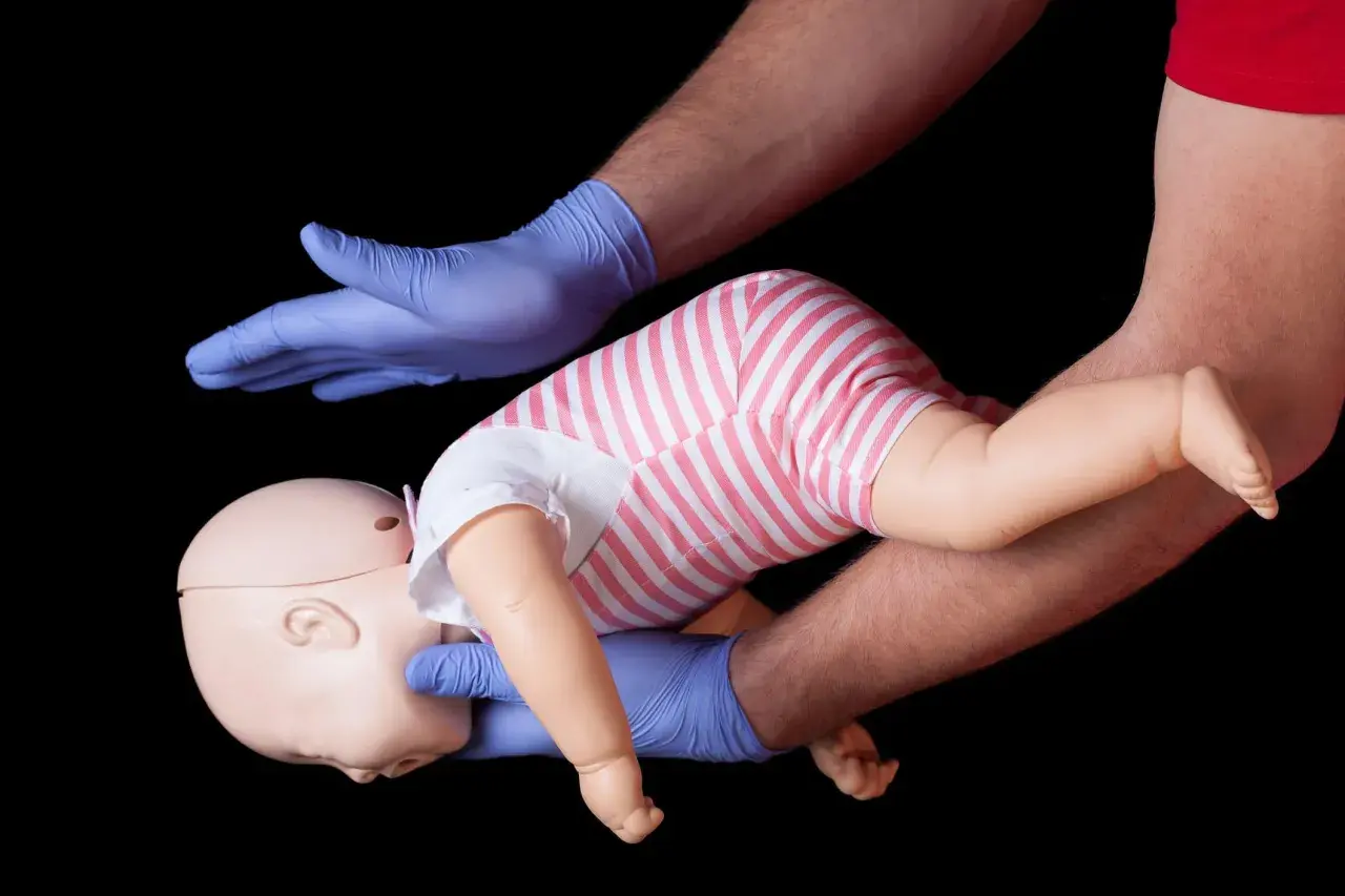 Esercitazione sulla manovra di Heimlich per bambini: mani guantate sostengono un manichino neonato, simulando un intervento di primo soccorso.