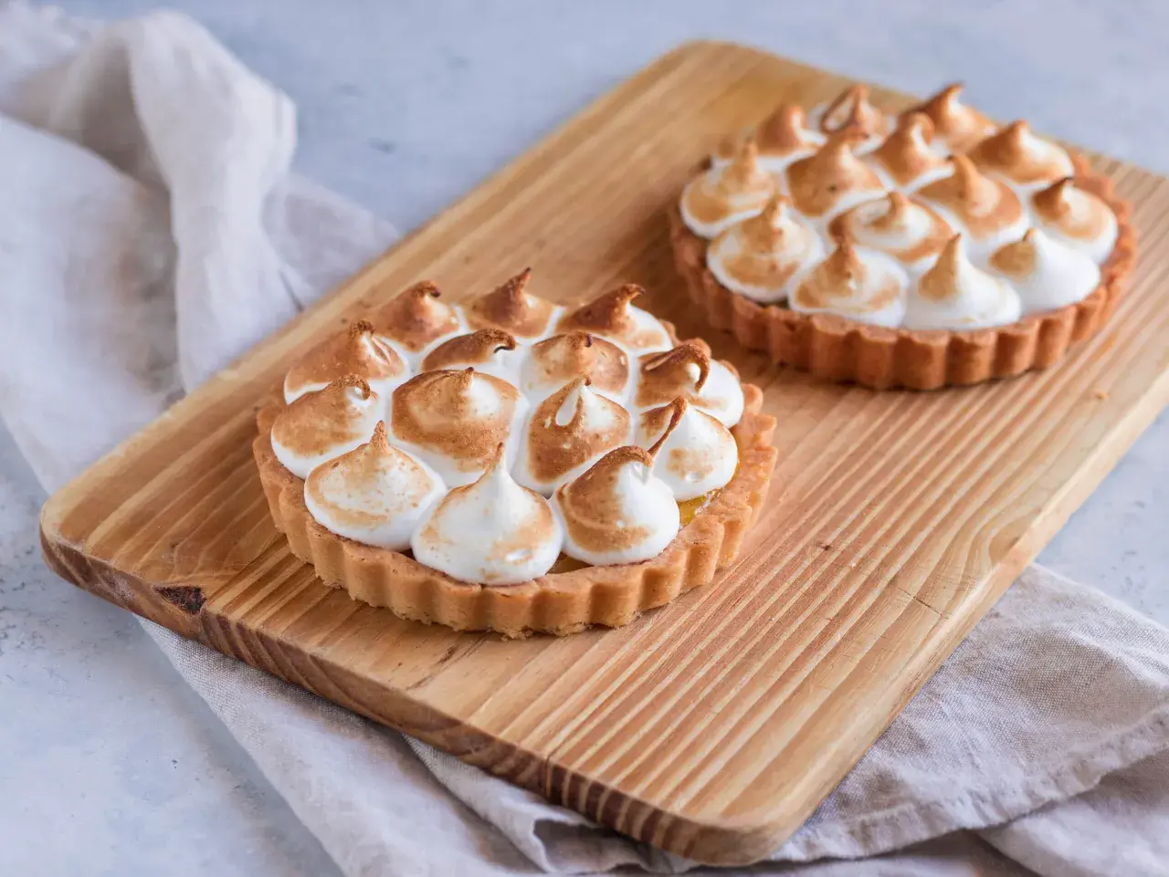 Due crostatine con crema al limone e meringa all'italiana, dorata e soffice, su un tagliere di legno.