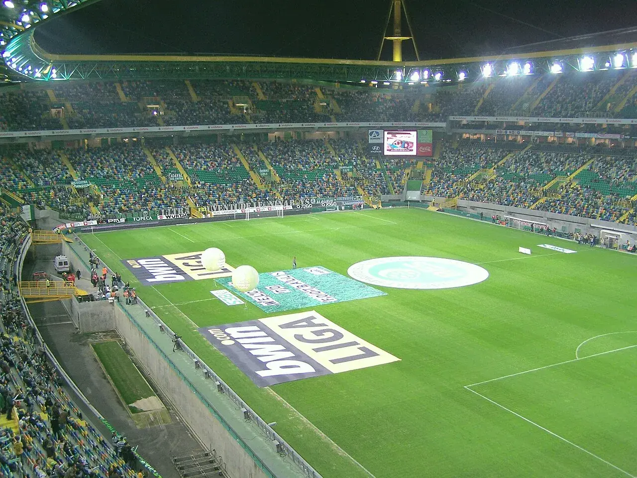 Widok na pełny stadion Sporting Lizbona, z zieloną murawą, trybunami wypełnionymi kibicami i reklamami na boisku.