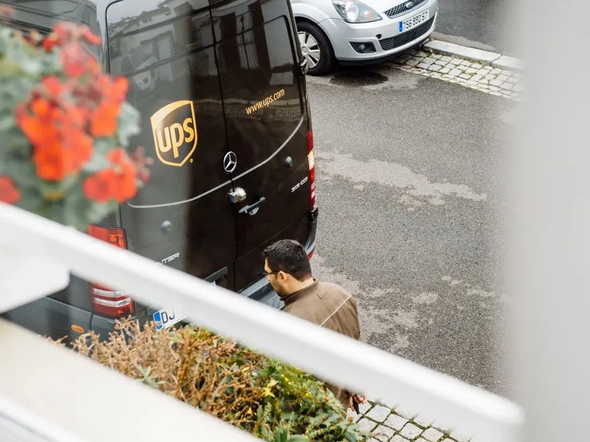 Kurier UPS z paczką zastanawia się, ups gdzie nadać. Obok stoi srebrny samochód.