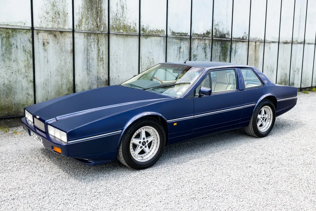 Granatowy Aston Martin Lagonda z lat 80. na tle industrialnej ściany.