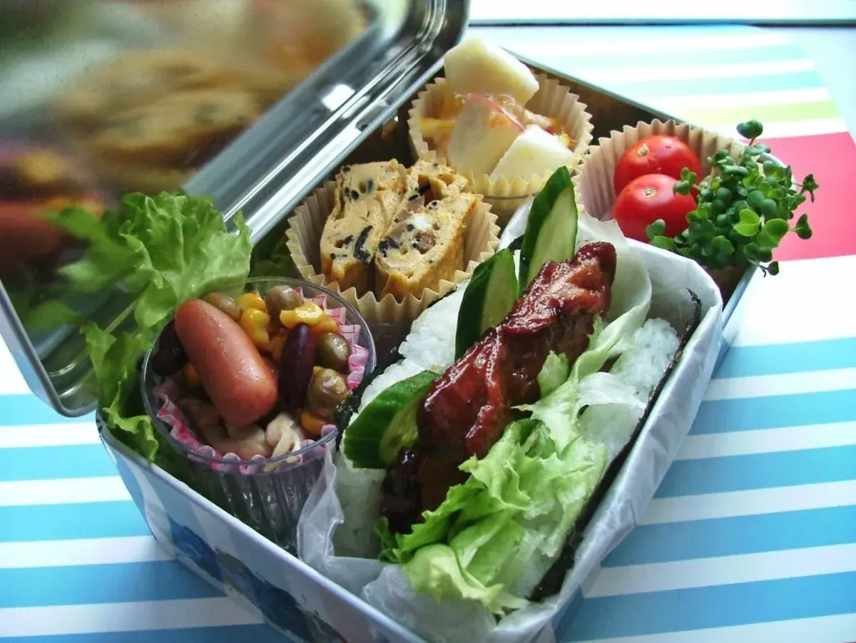 A delicious bento box with a glazed meat skewer, rice, lettuce, egg roll, sausage, beans, corn, tomatoes, and sprouts.