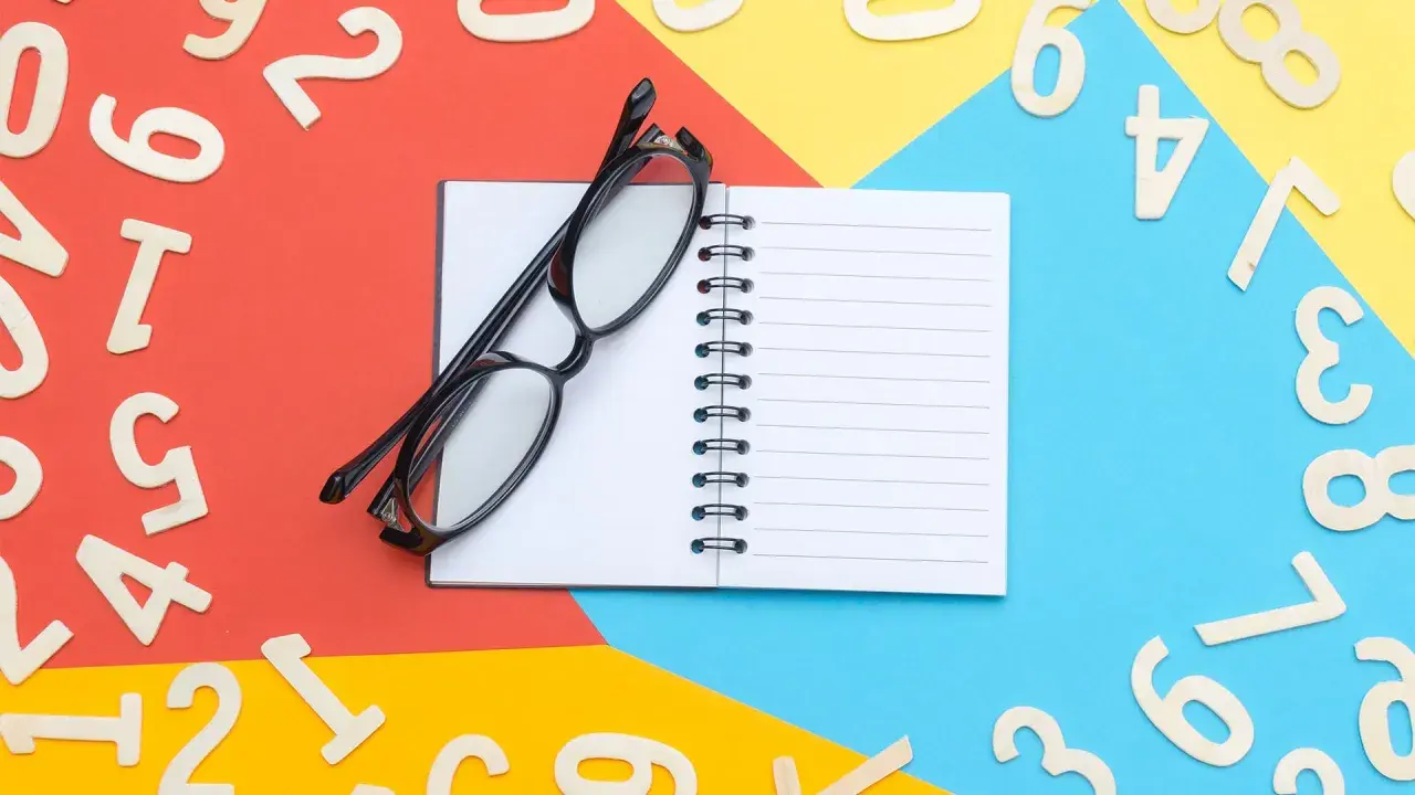Occhiali neri su un quaderno aperto, circondati da numeri in legno sparsi su sfondi colorati. Un promemoria visivo per imparare l'inglese, magari il numero 66.