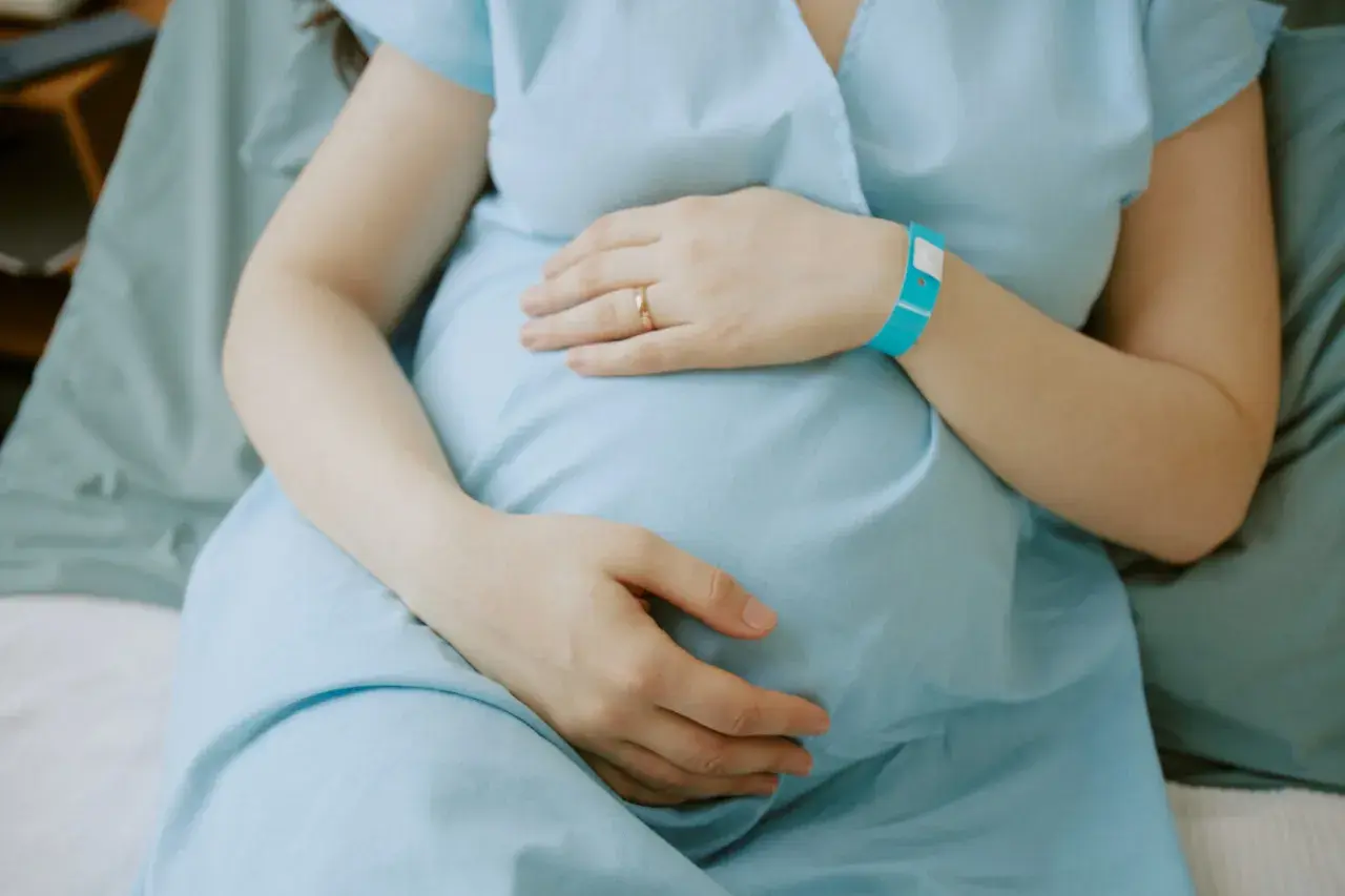Ciężarna kobieta w niebieskiej koszuli szpitalnej trzymająca się za brzuch, z opaską na nadgarstku.