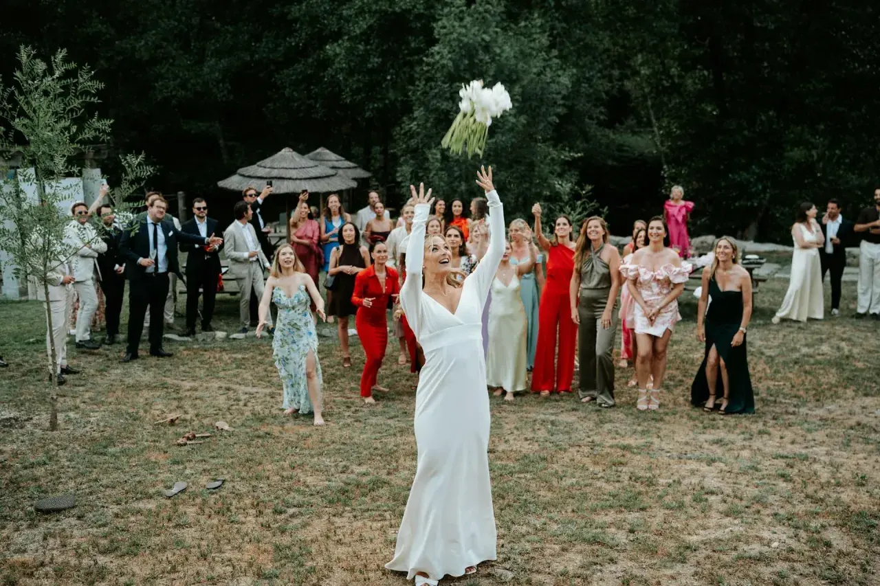 Panna młoda w białej sukni rzuca bukietem, a zgromadzeni goście, w tym panie w kolorowych kreacjach, czekają na jego złapanie. To piękny moment celebrujący tradycje weselne.