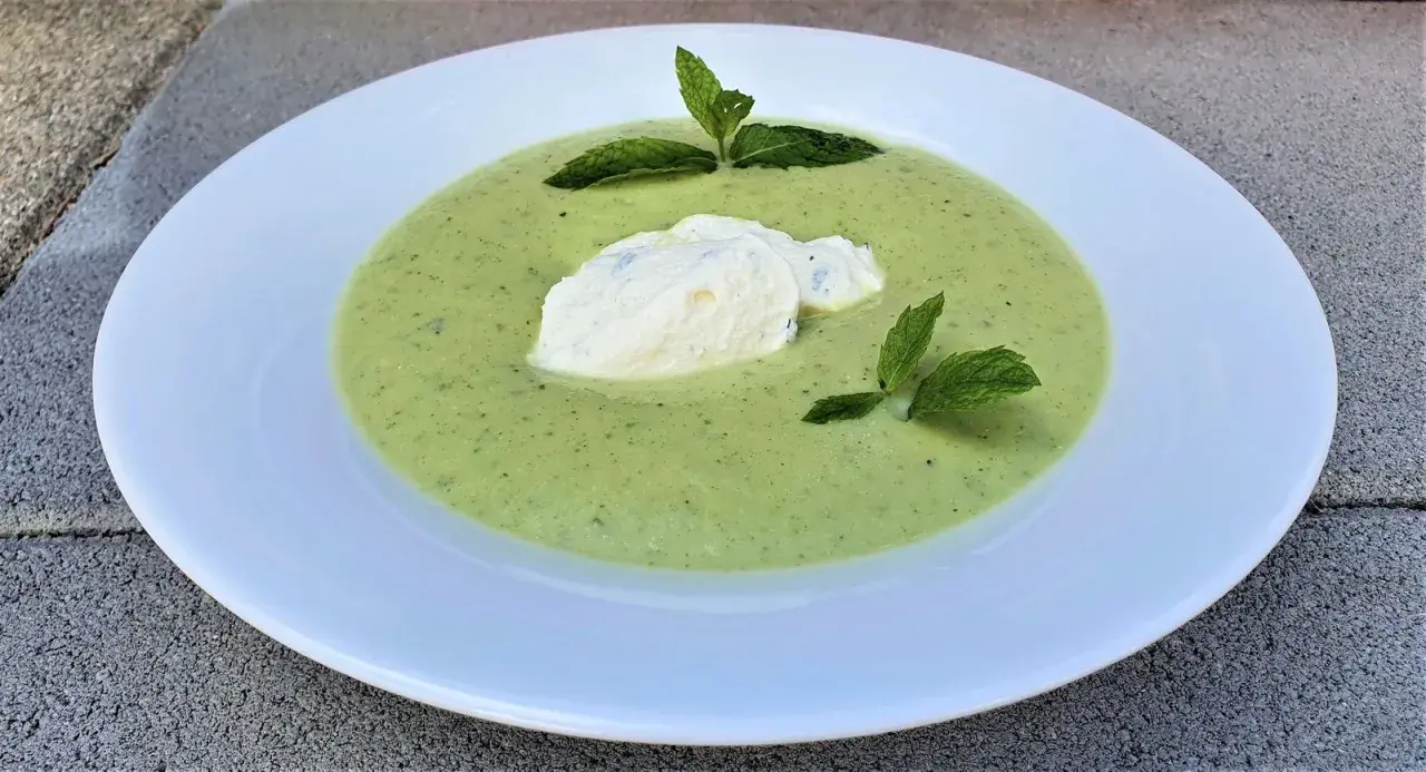 Velouté de courgette onctueux, garni d'une quenelle de fromage frais et de feuilles de menthe, servi dans une assiette blanche.