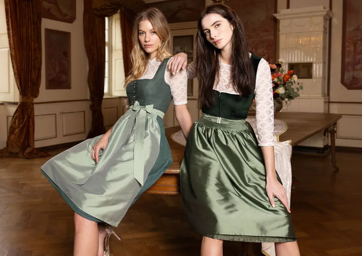 Zwei Frauen in Tracht. Die eine trägt ein grünes Dirndl mit einer hellgrünen Schürze, die andere ein dunkelgrünes Dirndl mit einer glänzenden, grünen Schürze. Eine dirndlschürze braun wäre eine schöne Ergänzung.