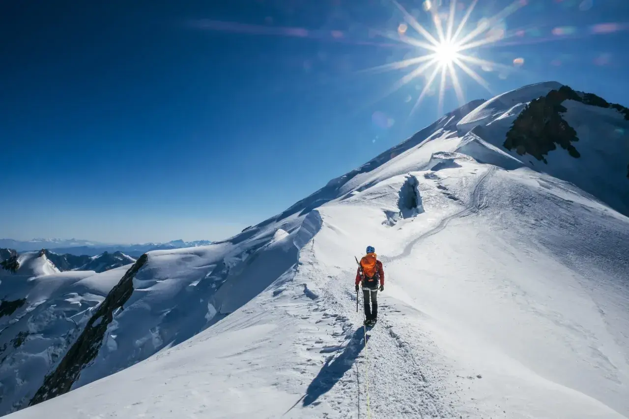 Un alpiniste solitaire progresse sur une crête enneigée, le soleil éclatant au-dessus du sommet du Mont-Blanc.