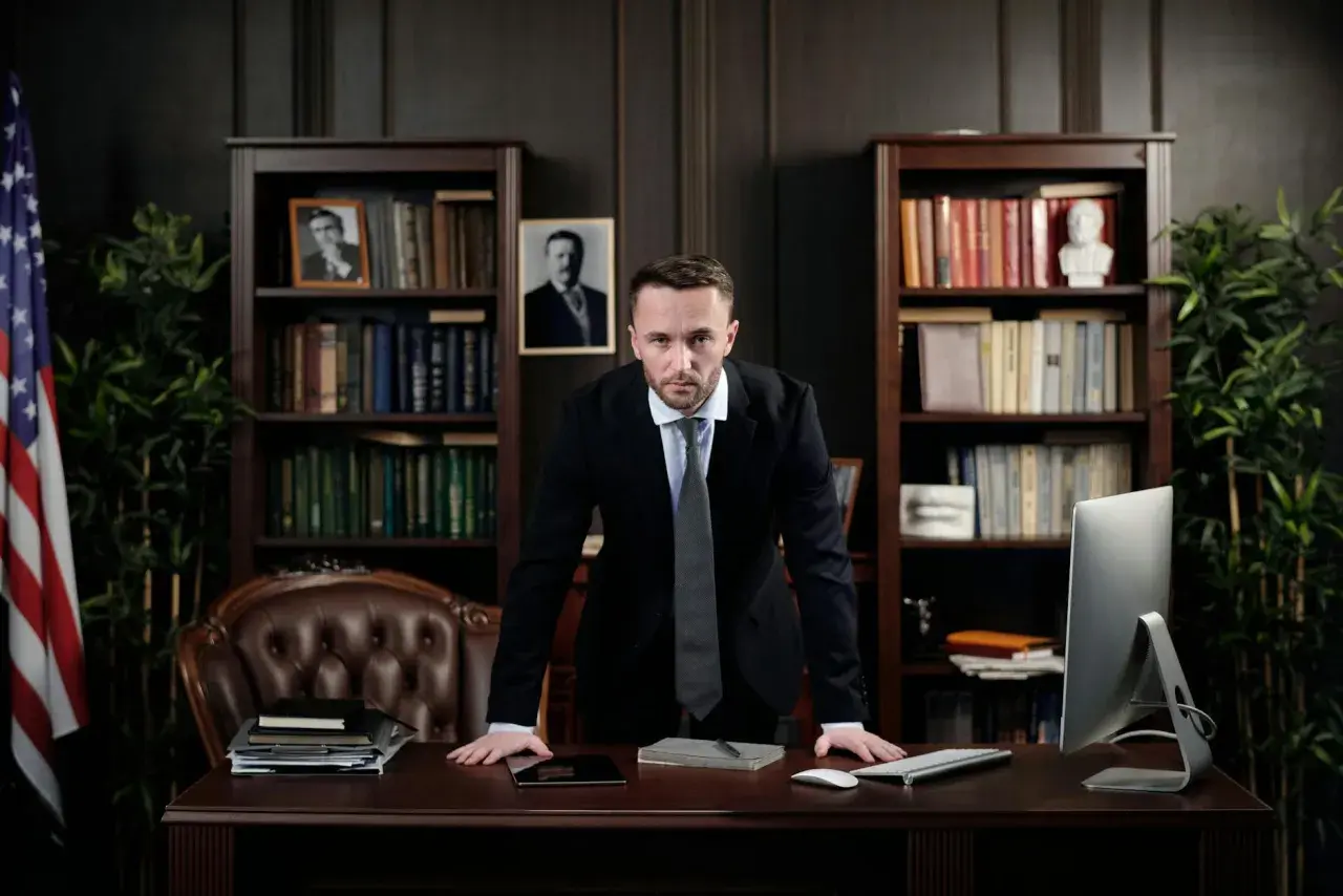 Un homme en costume se tient derrière un bureau, prêt à rendre un jugement contradictoire. Des livres et un drapeau américain ornent la pièce.