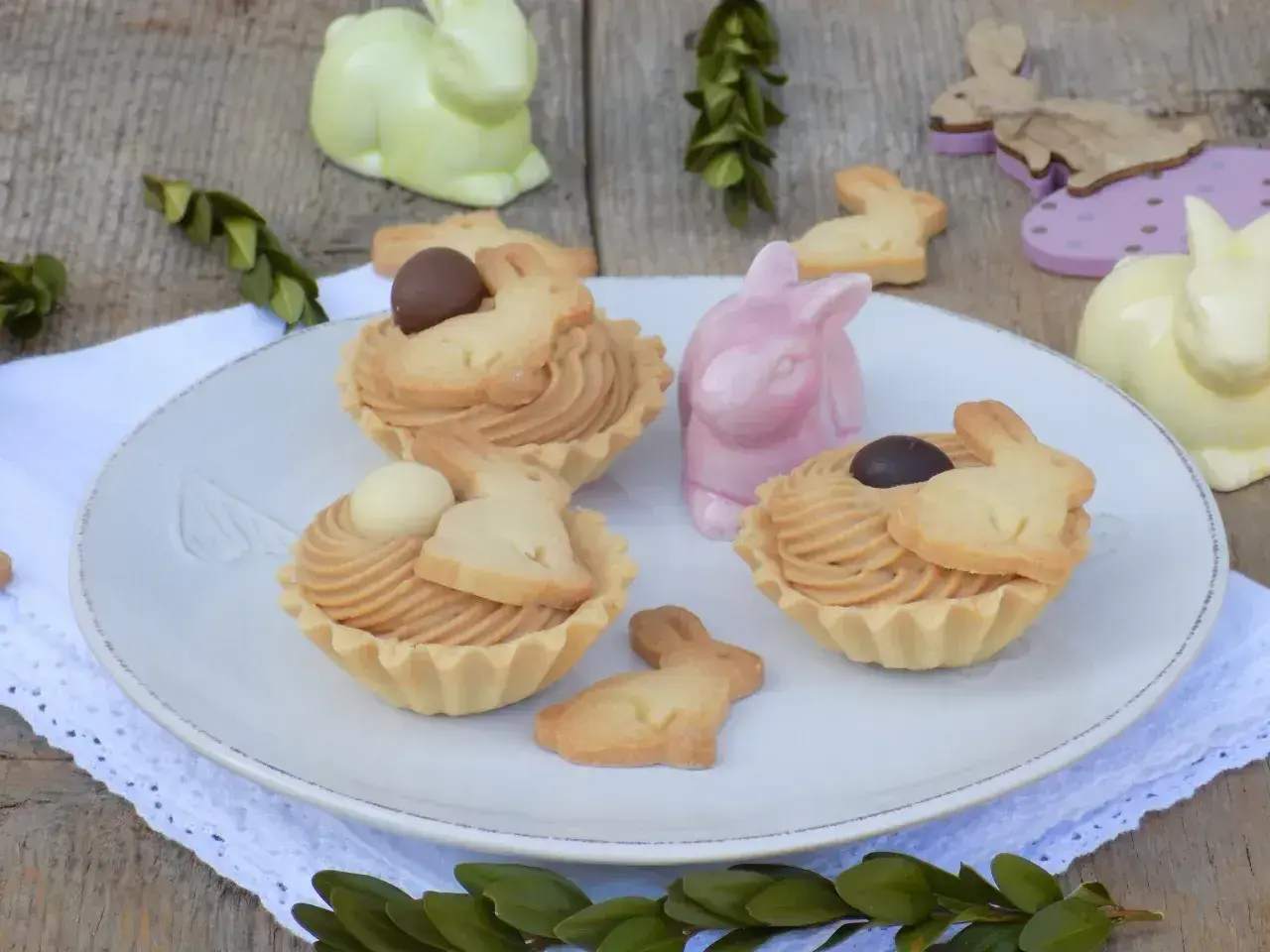 Wielkanocne babeczki z mascarpone, ozdobione ciasteczkami w kształcie króliczków i czekoladowymi jajeczkami, na talerzu wśród ceramicznych zajączków.