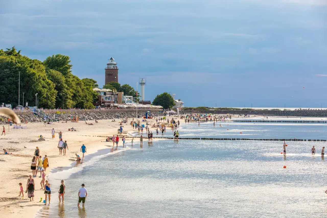 Tłumy ludzi na piaszczystej plaży, latarnia morska w tle. Idealne miejsce na tanie miejscowości nad morzem.