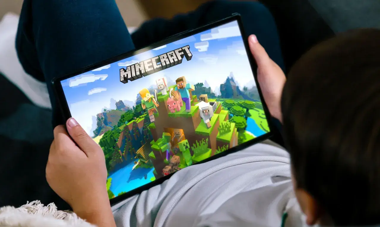 Dziecko gra na tablecie w Minecrafta, najpopularniejszą grę na świecie, eksplorując pikselowy świat z postaciami i zwierzętami.