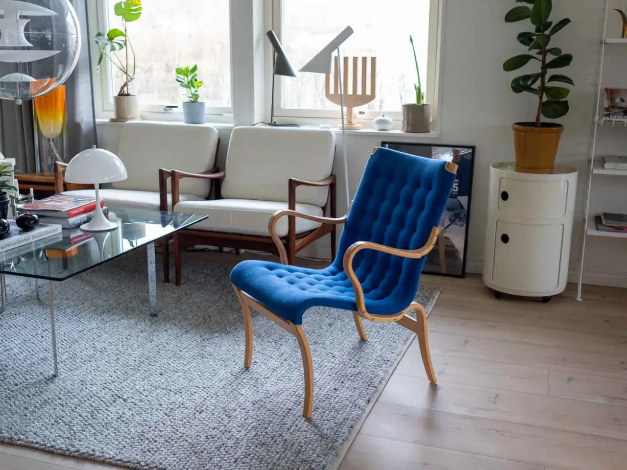 En blå Bruno Mathsson stol med böjda träarmstöd står på en grå matta. Bredvid syns vita soffor och ett glasbord.