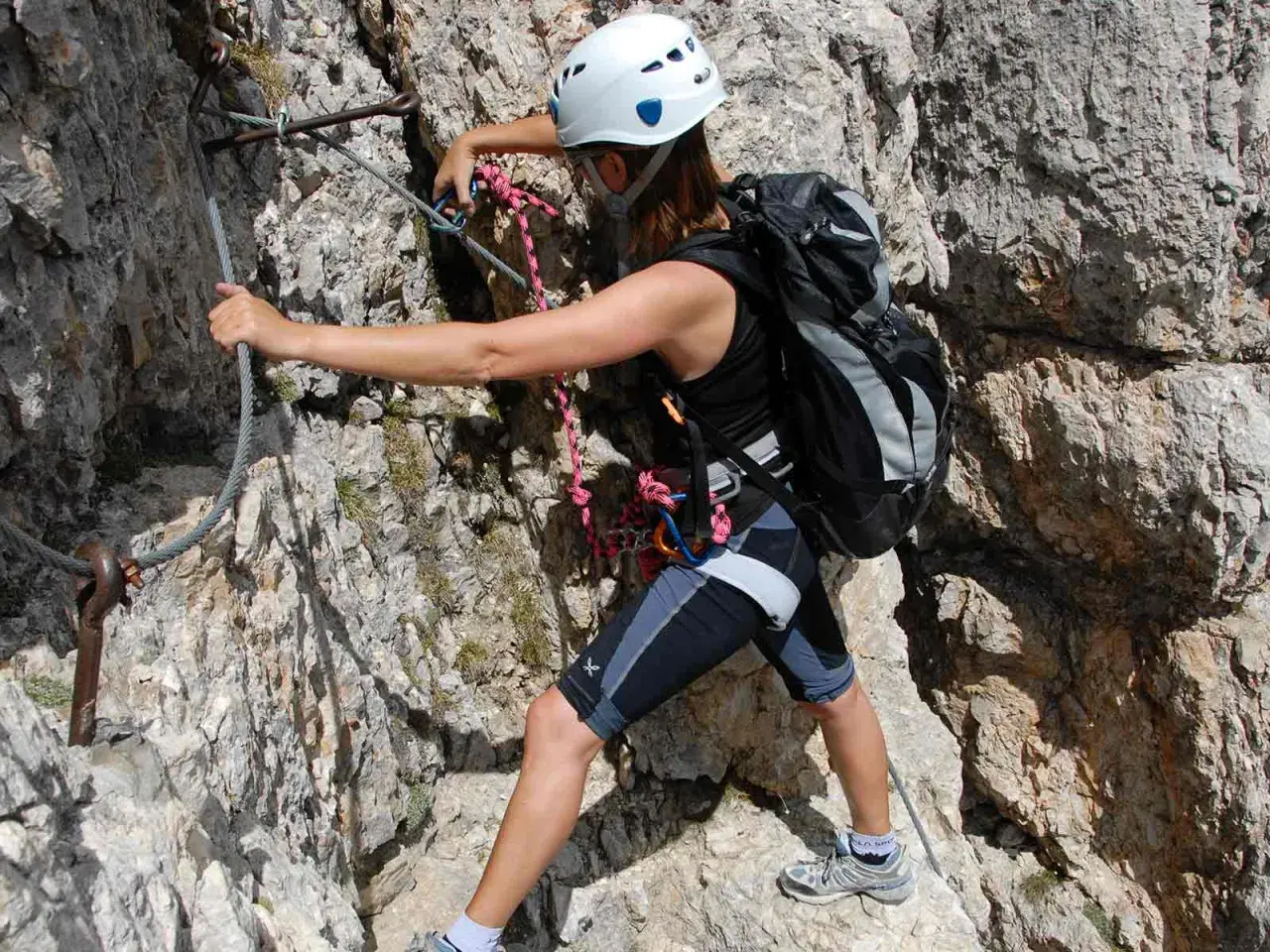 Escaladora con casco y mochila se asegura a un cable en una pared rocosa. La puntuación escalar de su equipo es alta.