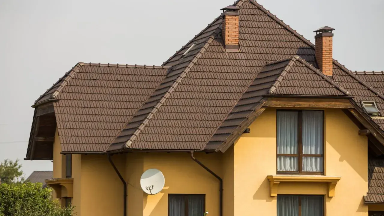 Dach naczółkowy konstrukcja z brązowej dachówki, z ceglanym kominem i anteną satelitarną na żółtej ścianie domu.