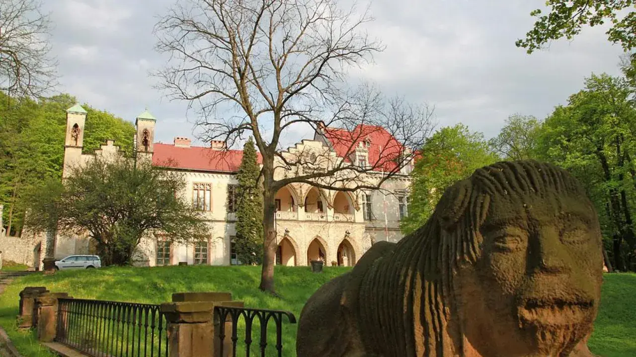 Kamienna rzeźba twarzy przed pałacem w Młoszowej. Budynek z czerwonym dachem i arkadowym gankiem otoczony zielenią.