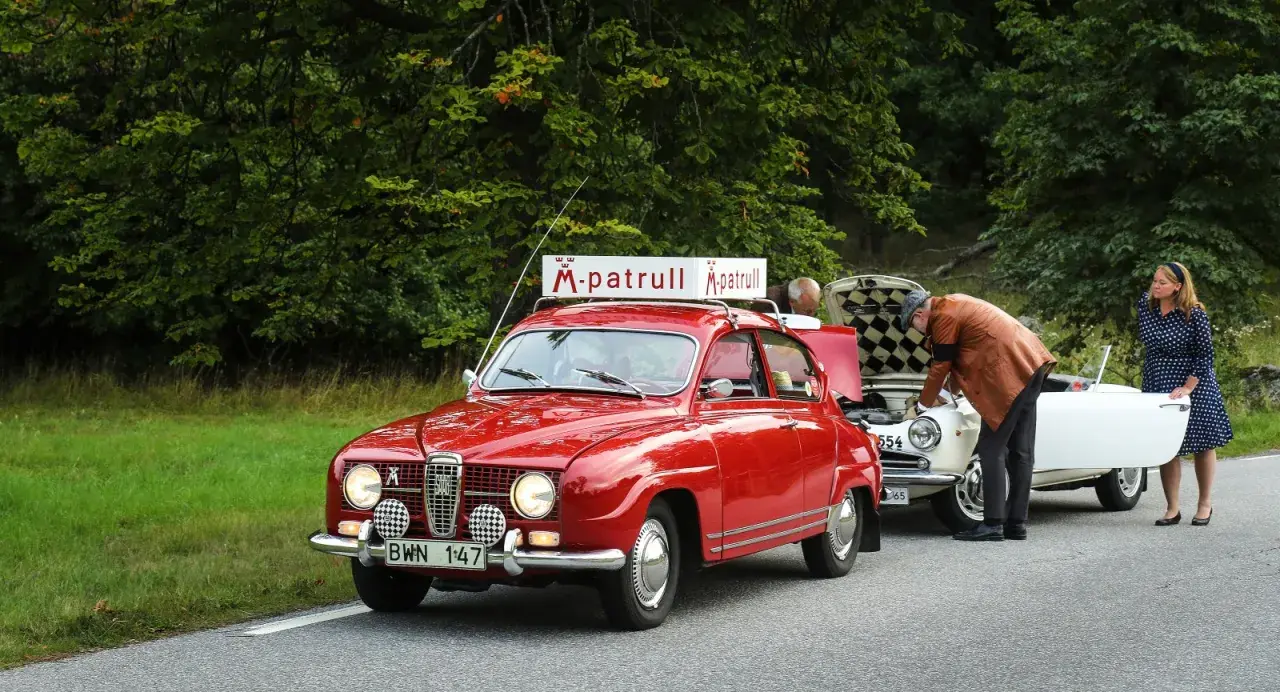 Röd Saab 96 V4 med "M-patrull" skylt på taket. En man tittar i motorhuven på en vit bil, en kvinna står bredvid.