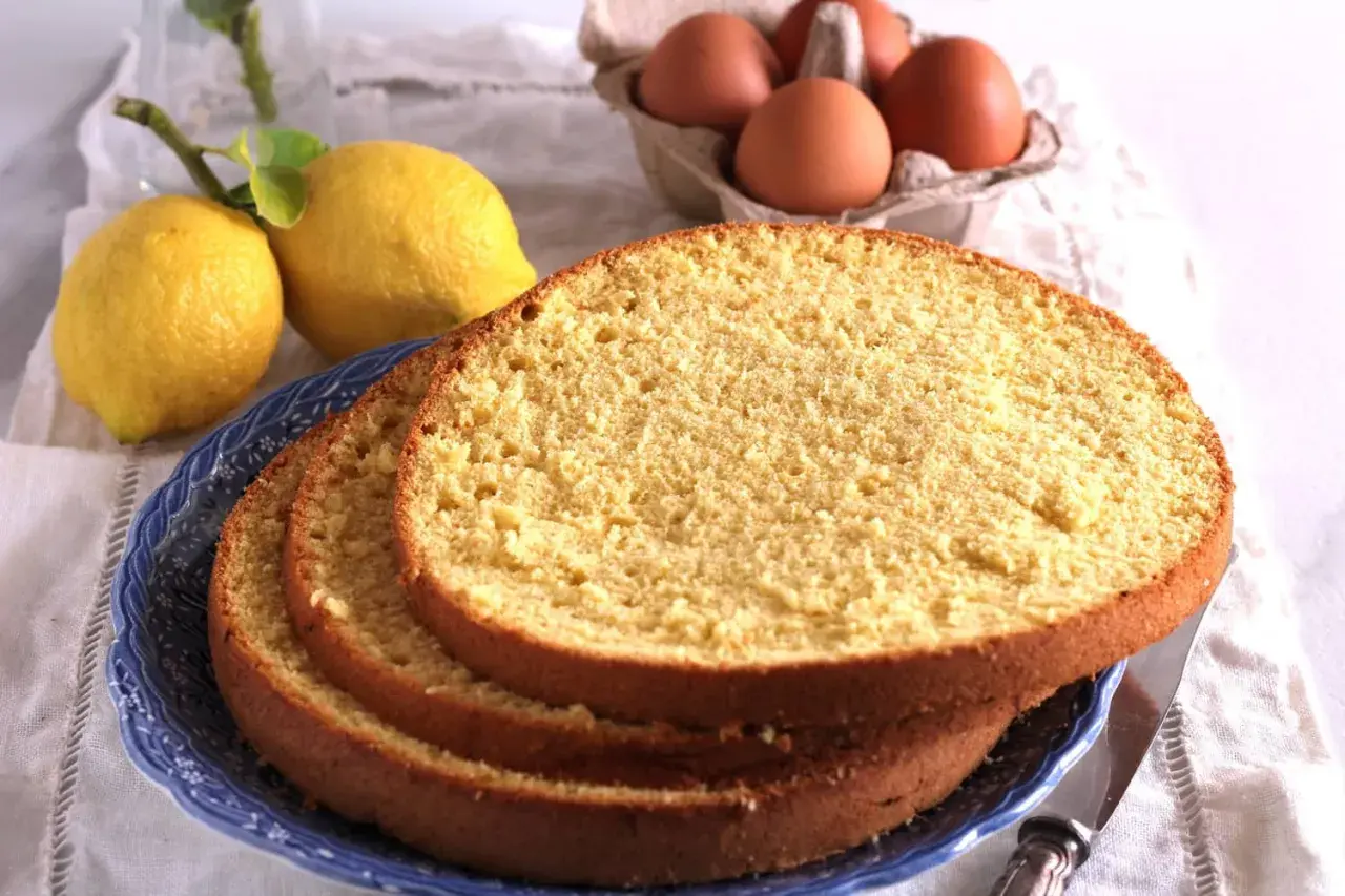 Fette di soffice sponge cake su un piatto blu, con limoni e uova sullo sfondo, pronte per una deliziosa torta.