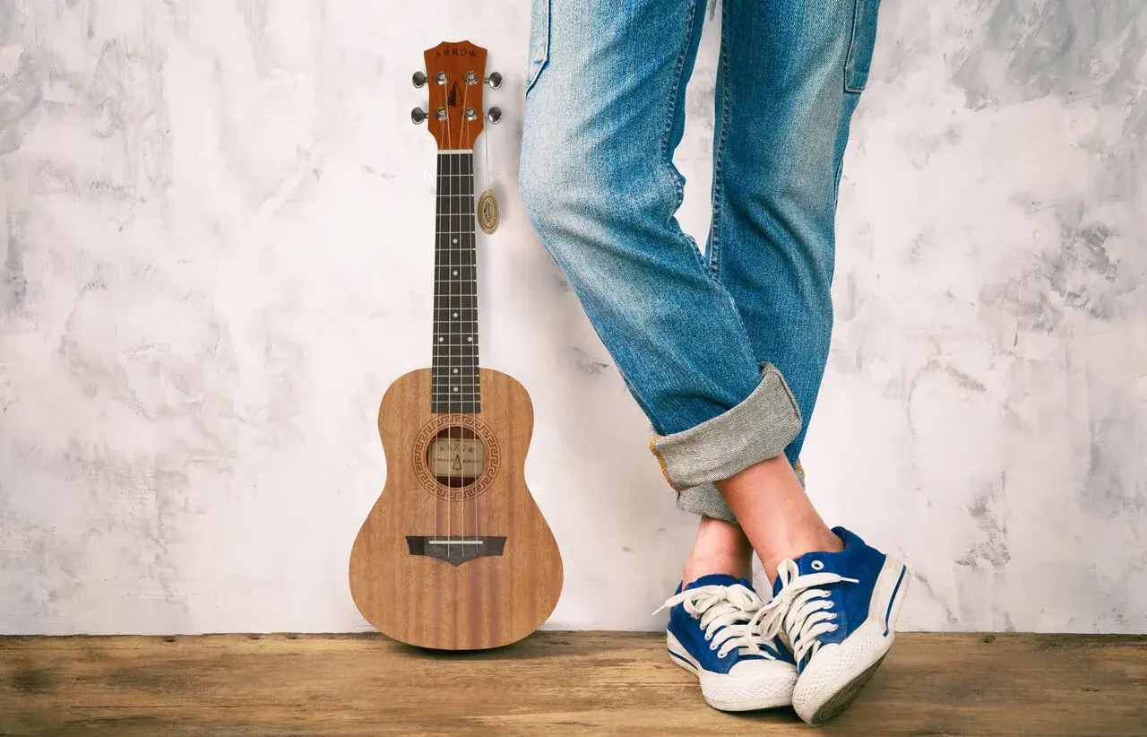 Drewniane ukulele obok osoby w jeansach i niebieskich trampkach. Idealne do grania letnich hitów na ukulele.