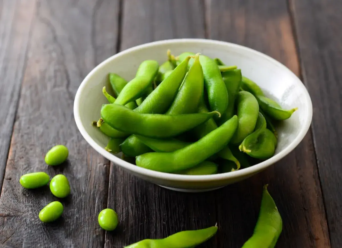 Miseczka pełna zielonej fasolki edamame na drewnianym tle. Kilka fasolek rozsypanych obok.