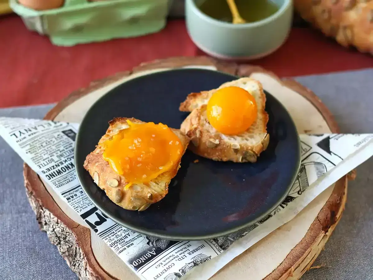 Sur une assiette, deux toasts de pain aux graines, l'un garni d'une texture orangée et l'autre d'un jaune d'œuf confit brillant.