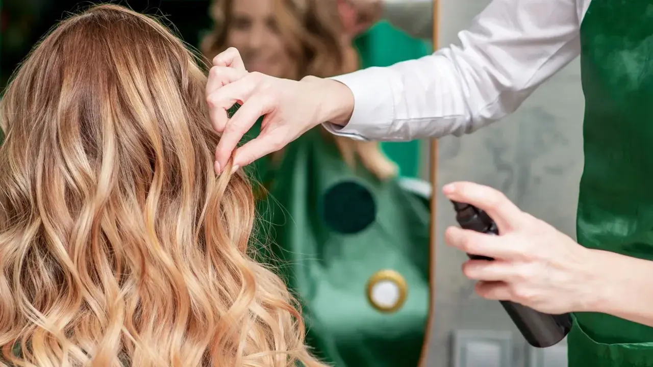 Fryzjerka utrwala fryzurę klientki, spryskując jej blond włosy lakierem. Perfekcyjny styling włosów.