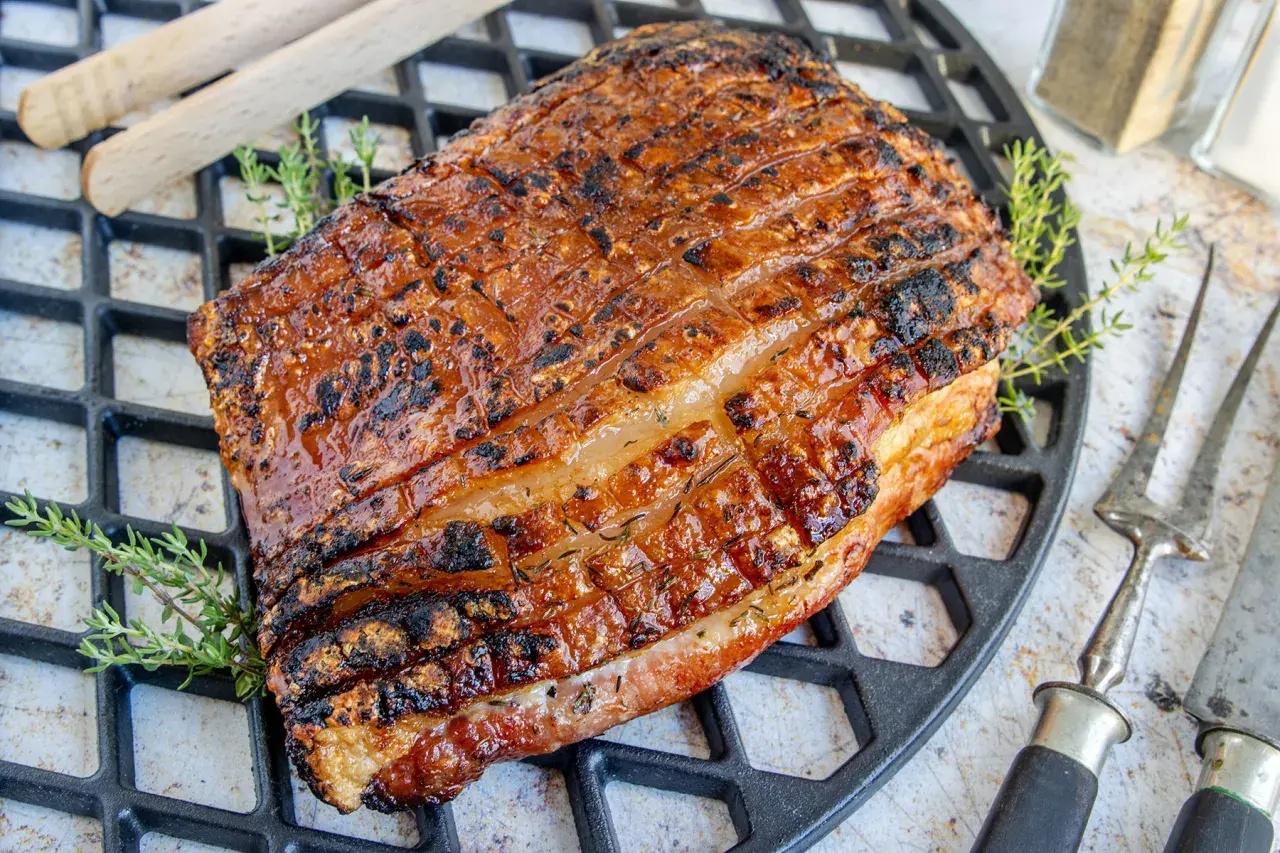 Perfekt gegrillter Schweinebauch mit knuspriger Schwarte liegt auf einem Grillrost. Kräuter und Grillbesteck deuten auf ein köstliches Mahl hin.