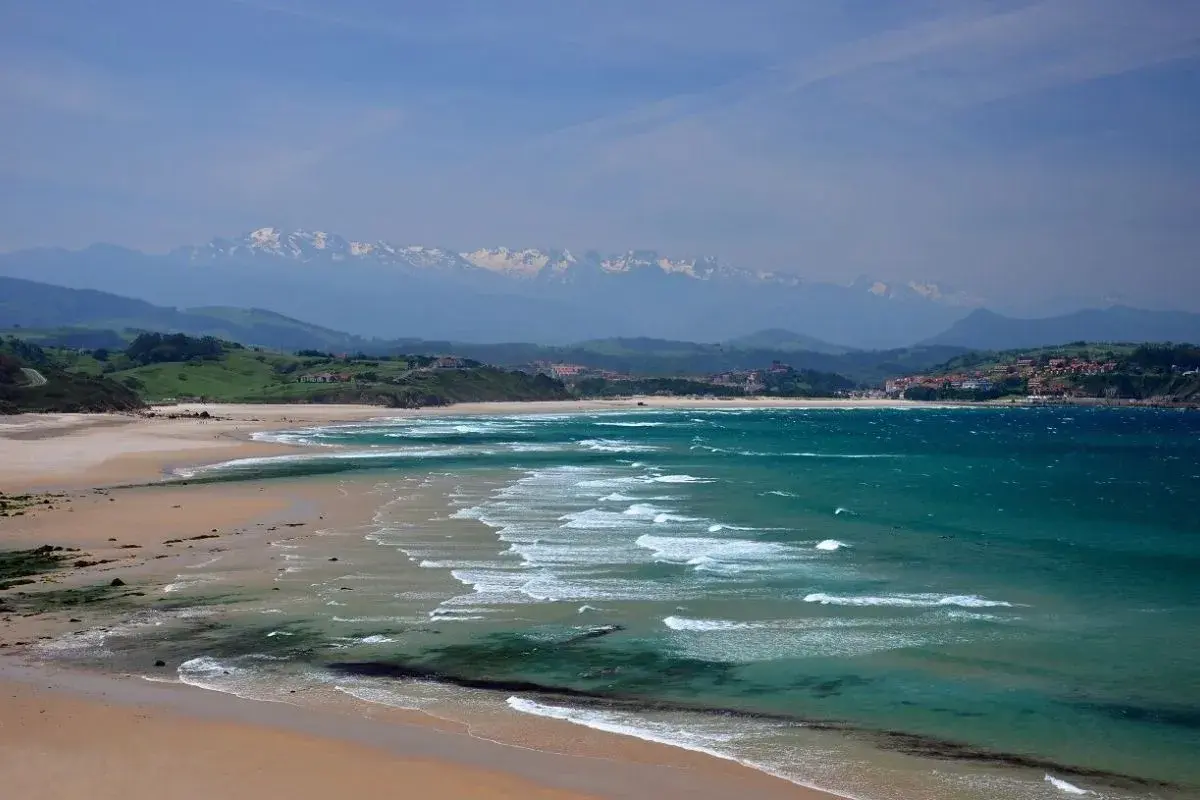 Amplias playas de arena dorada y aguas turquesas, con olas rompiendo suavemente. Al fondo, colinas verdes y montañas nevadas, un paisaje idílico de playas cerca de Comillas.
