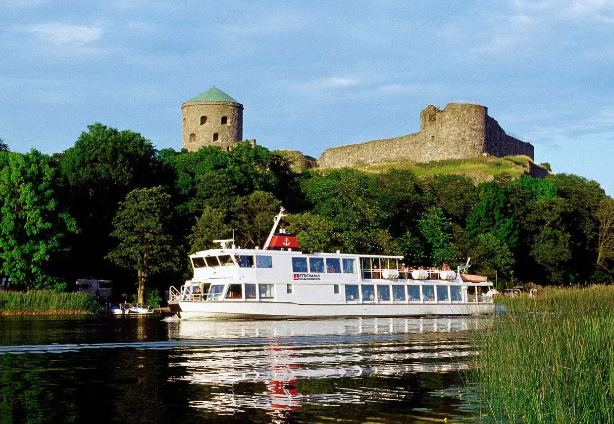 En vit båt från Strömma Boat Tours glider fram på vattnet, förbi grönskande träd och en gammal borg.