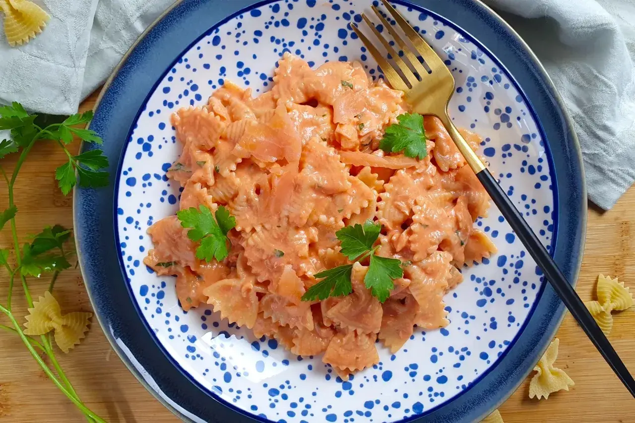Farfalle al salmone cremose, guarnite con prezzemolo fresco, servite su un piatto blu e bianco con una forchetta dorata.