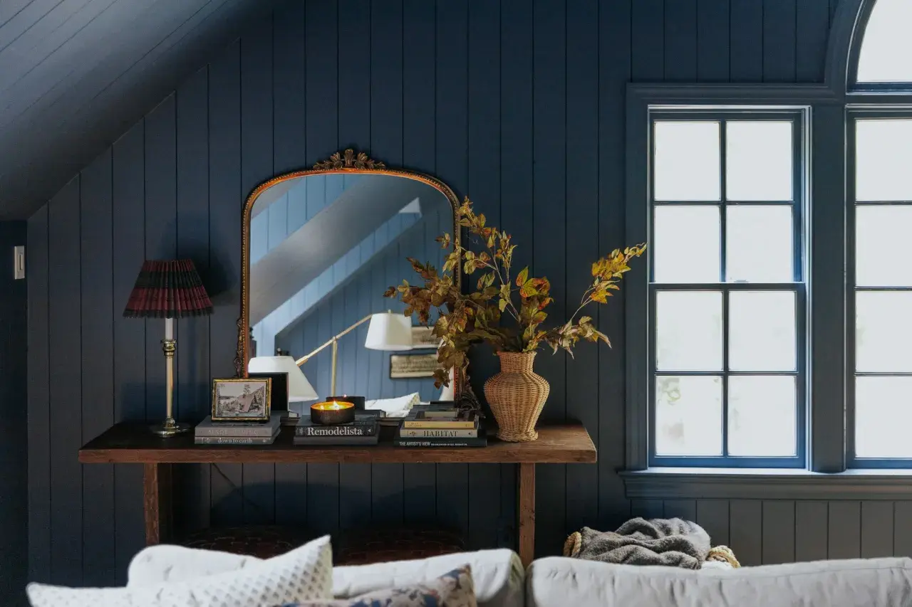 A rustic style console table with a lamp, books, and a vase of autumn leaves sits against a dark blue paneled wall, reflecting in an ornate mirror.