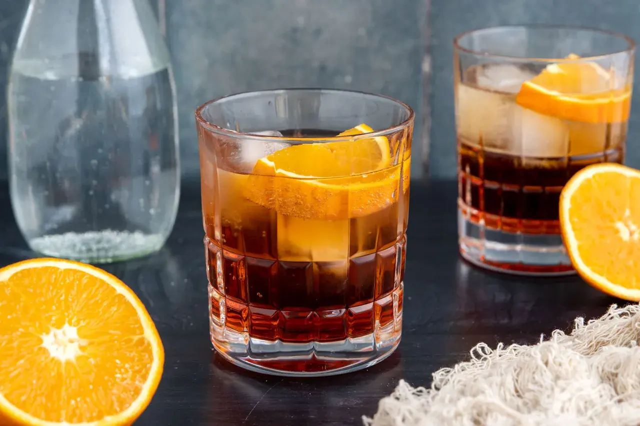 Ein erfrischender Americano Cocktail mit Eis und Orangenscheiben, daneben eine Karaffe Wasser und frische Orangen.