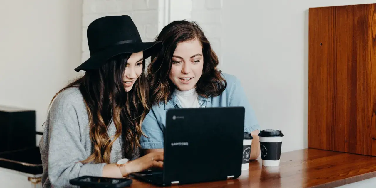 Två unga kvinnor diskuterar sin skuldkvot och bolån framför en laptop, med kaffekoppar på bordet.