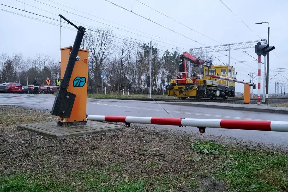 Żółta lokomotywa z dźwigiem zbliża się do przejazdu kolejowego bez zapór. Na pierwszym planie pomarańczowy słup z podniesionym ramieniem.