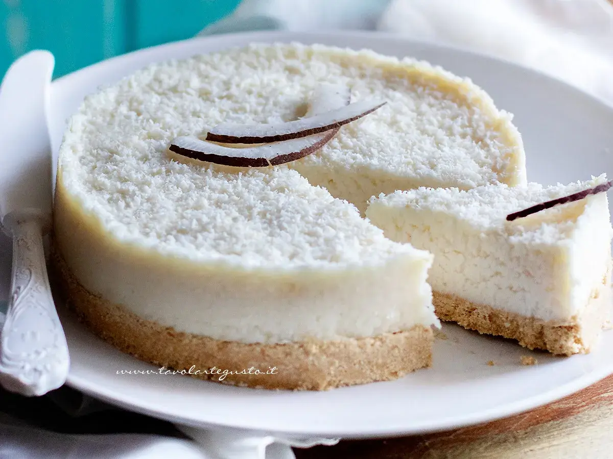 Delizioso cheesecake al cocco, ricoperto di cocco grattugiato e decorato con scaglie di cocco fresco. Una fetta è già stata tagliata, invitando a gustare questo dolce tropicale.