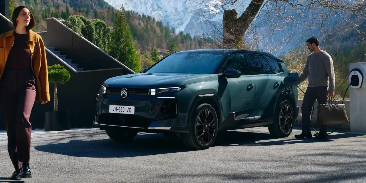Nowy Citroën C5 Aircross 2025, zielony SUV, ładowany przy górskim domu. Kobieta w płaszczu wychodzi z budynku.