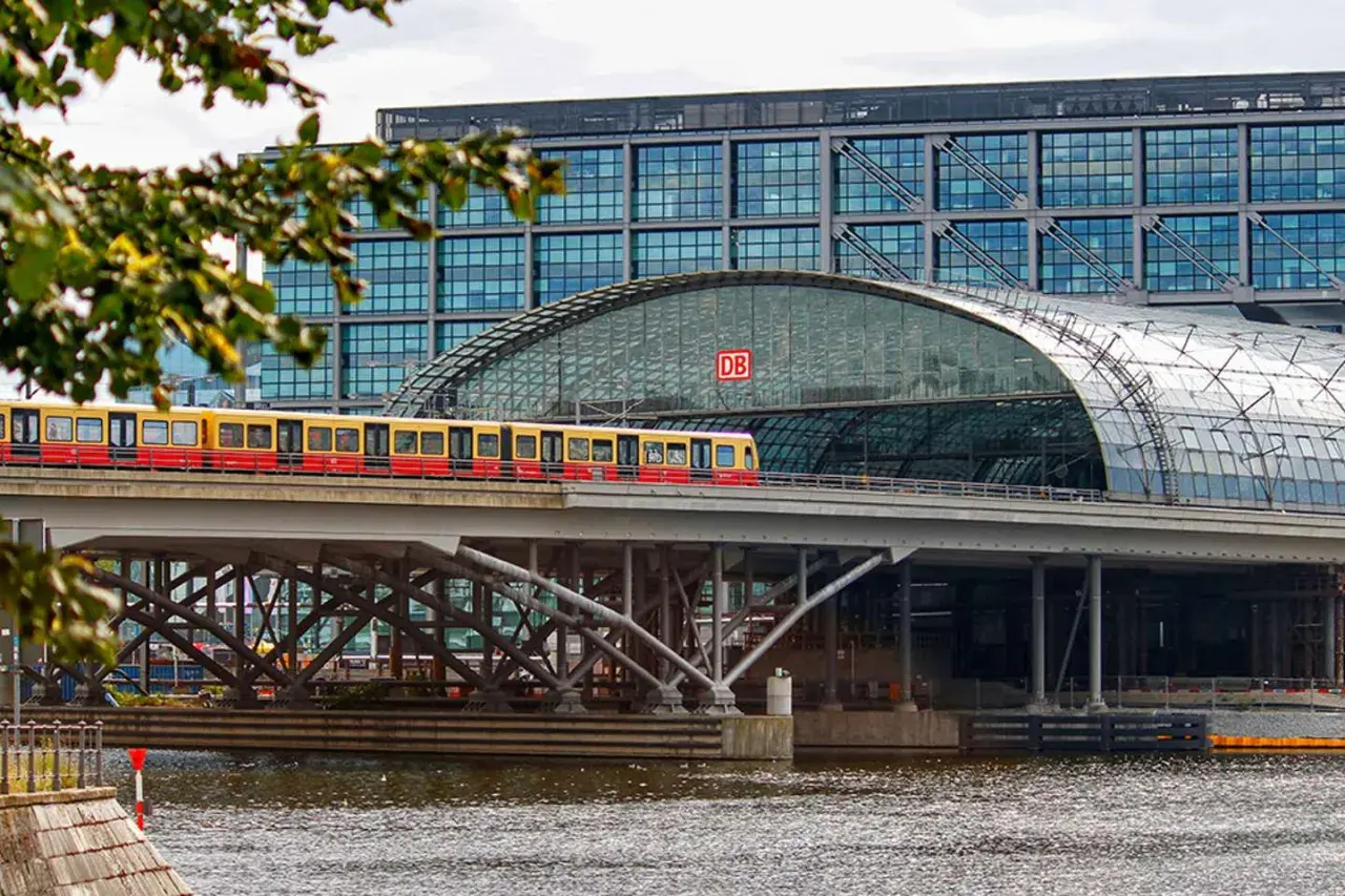 Ein roter und gelber Zug fährt über eine Brücke vor dem Berliner Hauptbahnhof. Die Verbindung Berlin Dresden ist hier gut sichtbar.