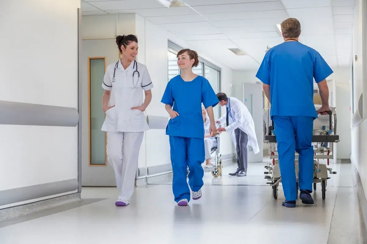 In un corridoio ospedaliero, personale medico si muove con urgenza. Un uomo spinge una barella, mentre due infermiere camminano velocemente, forse a seguito di un evento avverso.