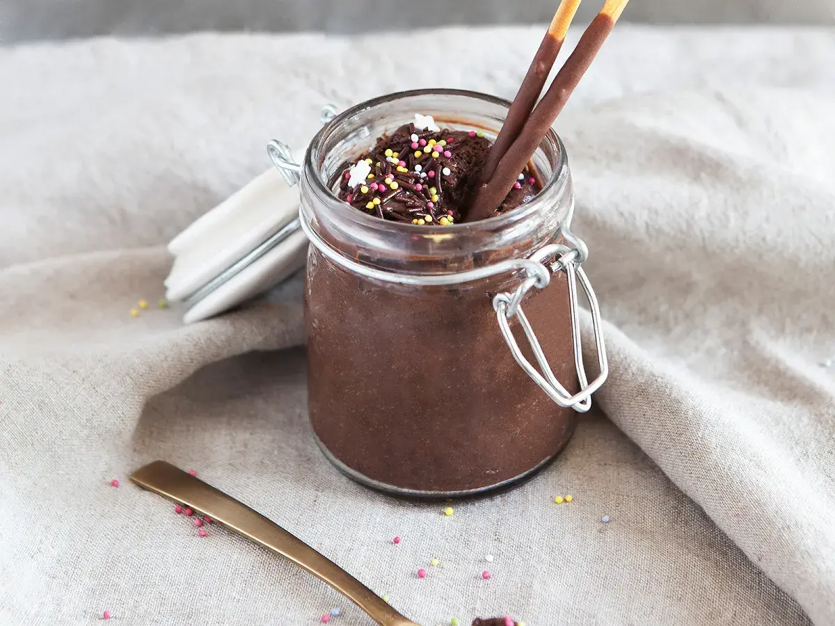 Un pot de mousse au chocolat mascarpone, garni de vermicelles colorés et de bâtonnets au chocolat. Une cuillère dorée repose à côté.
