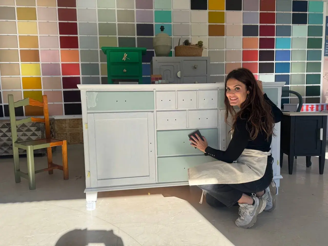 Mujer lijando un armario en proceso de pintura, con un fondo de muestras de colores.