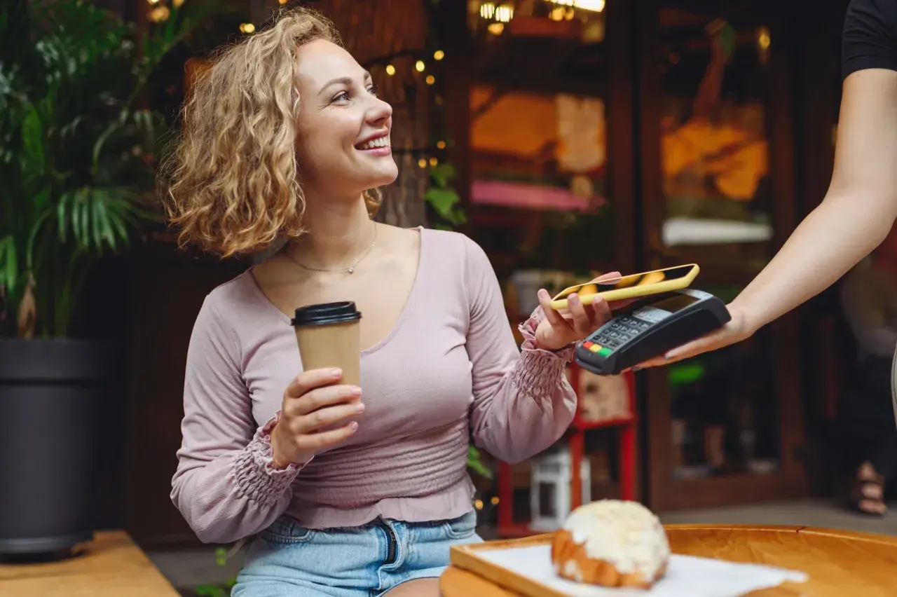 Uśmiechnięta kobieta z kawą płaci telefonem zbliżeniowo za croissant. Szybki przelew na telefon w kawiarni.