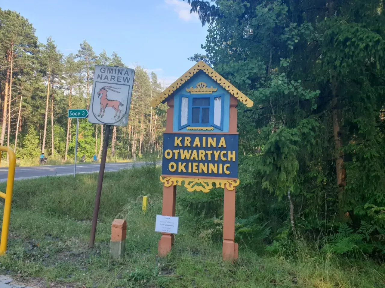 Drewniana tablica wita w Krainie Otwartych Okiennic. Obok znak Gminy Narew.
